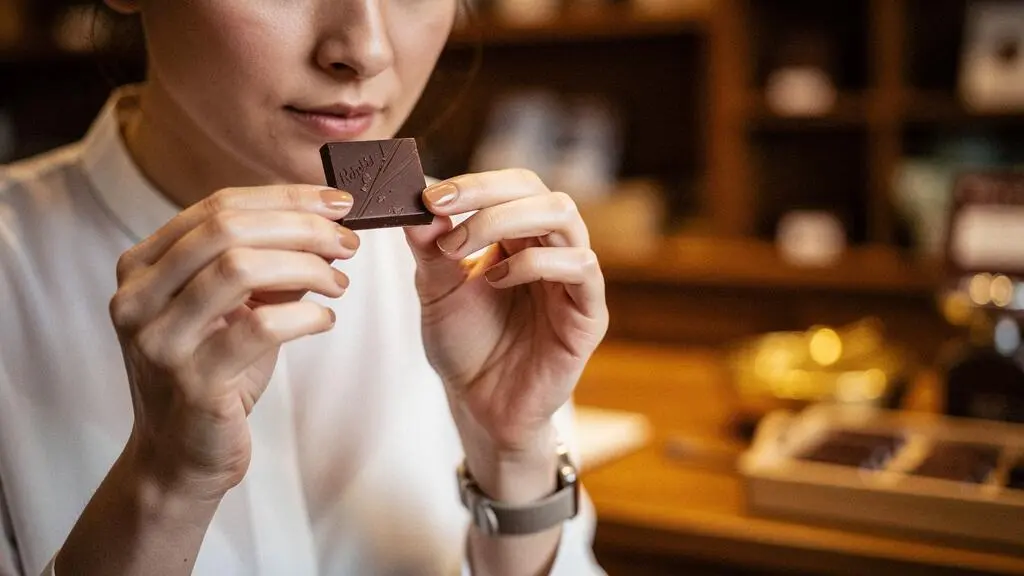 Gros plan élégant de mains tenant un carré de chocolat noir près du nez pour une dégustation raffinée, dans une ambiance feutrée à la française