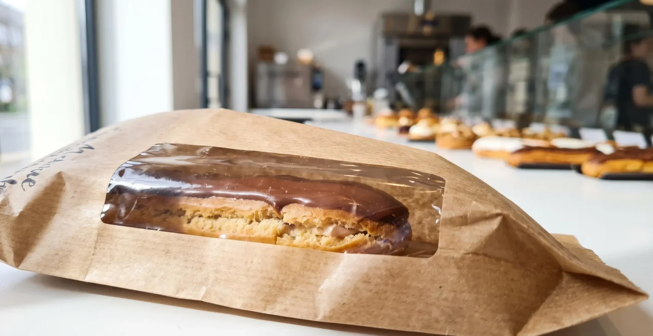 Gros plan détaillé sur un sachet kraft avec fenêtre transparente laissant voir un éclair au chocolat à l'intérieur, texture du papier visible